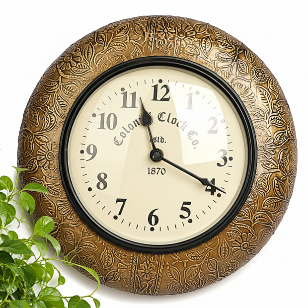 An ornate round wall clock with a textured bronze-brown floral frame and a cream-colored face, shown against a white background with green foliage in the lower left corner.